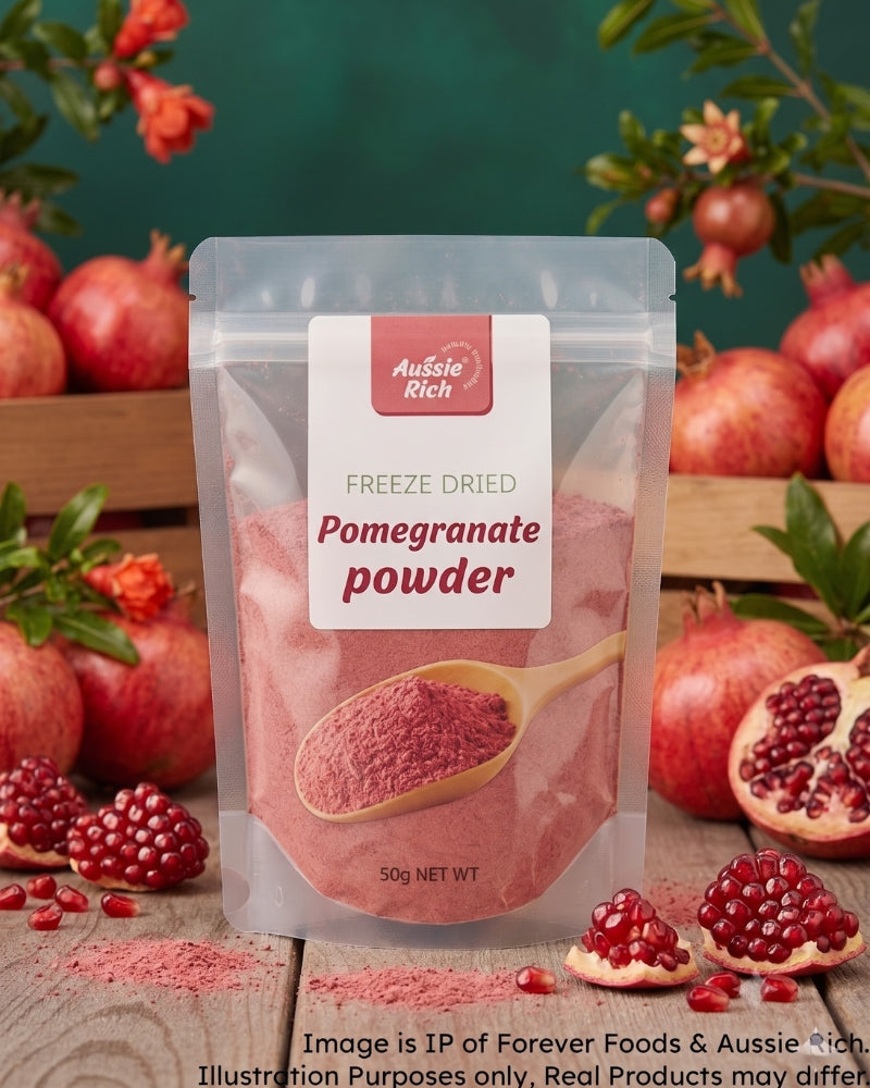 A 50g stand-up pouch of Aussie Rich Freeze Dried Pomegranate Powder, displayed on a rustic wooden surface surrounded by fresh pomegranates and green leaves against a dark green background.