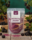 A stand-up pouch of Aussie Rich Freeze Dried Blackberry Powder, surrounded by fresh blackberries, green leaves, and wooden crates on a rustic wooden surface against a dark green background.
