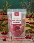 A stand-up pouch of Aussie Rich Freeze Dried Sour Cherry Powder, displayed on a rustic wooden surface surrounded by fresh cherries and green leaves against a dark green background.