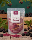 A stand-up pouch of Aussie Rich Freeze Dried Boysenberry Powder, shown with a wooden spoon filled with the powder. The pouch is surrounded by fresh blackberries, green leaves, and scattered loose powder on a rustic wooden surface, with a dark green background.